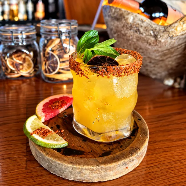 Cóctel amarillo en vaso escarchado con chile, menta y rodajas de cítricos